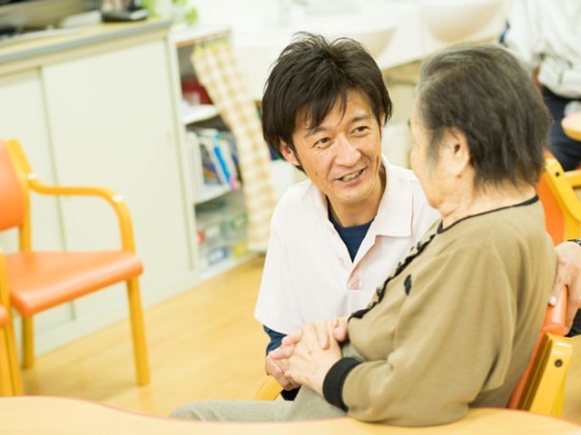 笑顔で会話するスタッフと利用者 スタッフイメージ スタッフが利用者の手を優しく握り、心温まる会話を交わしている様子です。安心のケアを感じていただけます。