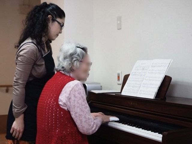 ピアノを弾く女性と支援者 ピアノ スタッフの優しいサポートのもと、ピアノ演奏を楽しむ姿が見られます。音楽を通したコミュニケーションで温かい雰囲気が育まれています。