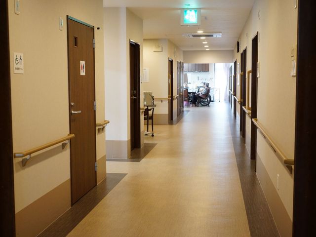 廊下と共用スペースの様子 廊下 すっきりとした廊下の先には、車椅子で会話を楽しむ方々が見えます。手すりが設置されており、安全に移動できる環境が整っています。