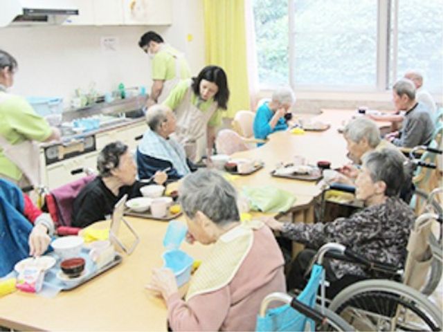 利用者の食事風景 日常 利用者たちが明るいダイニングで食事をとっています。スタッフの方々が丁寧にサポートしており、和やかな雰囲気が感じられます。