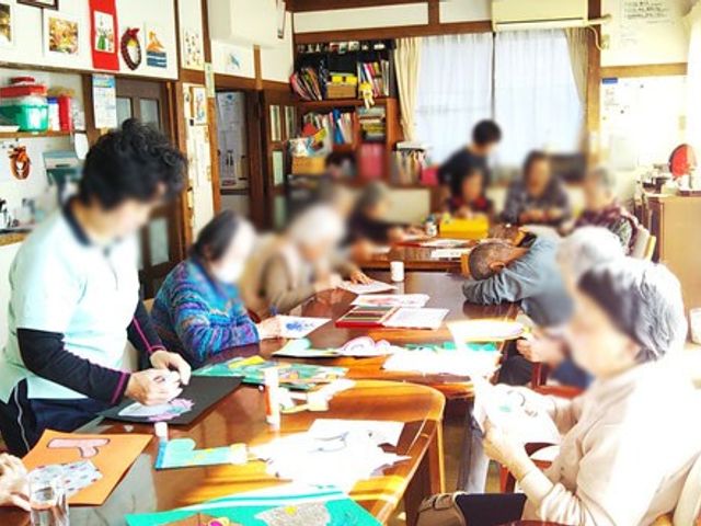共同作業中の部屋 多目的室 みんなでテーブルを囲み、手作業を楽しんでいます。明るい自然光が差し込む心地よい空間です。