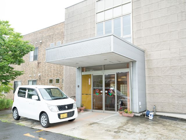 快適な施設のエントランス 外観 広々とした正面入り口には、花の鉢植えが飾られていて、迎え入れる雰囲気が漂っています。利用者の外出支援に活用されるような専用車両が停まっている様子から、日々の暮らしに必要な配慮が行き届いていることが伺えます。