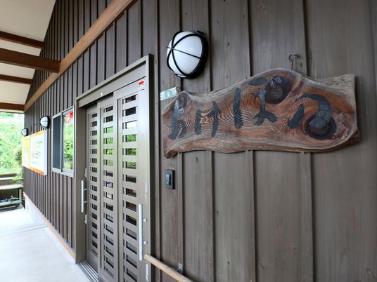 木の節のある一枚板に、墨で施設名称が記載された看板である。入口扉の横にインターホンや外灯とともに設置されている。
