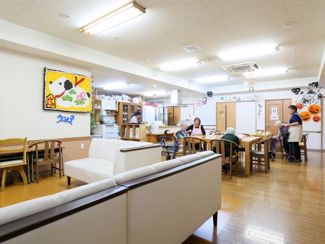 広々とした明るい空間 共有スペース ゆったり配置されたテーブルと椅子、心地よい光が差し込む食堂では、ケアを必要とする方々が日々の食事を楽しんでいます。白いソファが置かれ、居心地の良い待合スペースとしても機能しております。