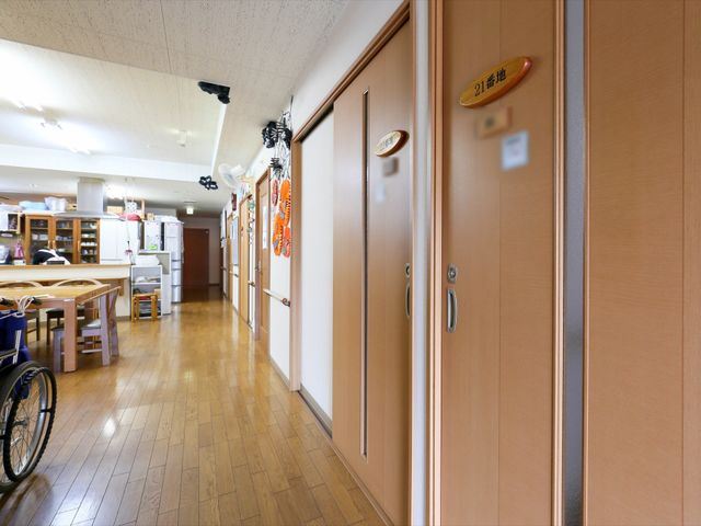 廊下と居室のドア 共有スペース ゆったりとした廊下には温かみのある木目のドアが並び、清潔感のあふれる明るい空間が広がっています。車椅子が置いてあり、バリアフリーに配慮された設計です。