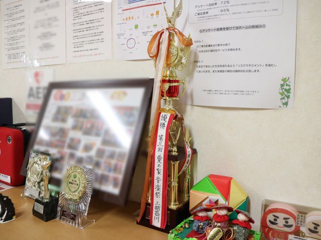 賞状とトロフィーの飾り棚 置き物 壁には表彰状が掲示され、努力と成果を称えるトロフィーや飾りがあり、職員の努力が感じられる光景です。