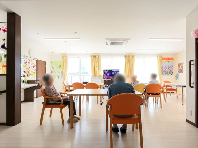 明るい共有スペースの風景 共有スペース 落ち着いた雰囲気の食堂で、利用者はテレビを視聴ながらくつろいでいます。おしゃれな壁の装飾も心地よい空間を演出しています。
