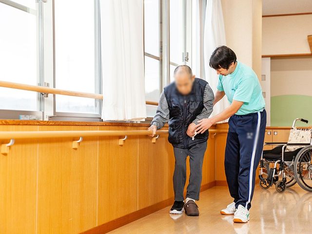 支援中の訓練室 機能訓練室 明るい室内で、介護職員が利用者を優しくサポートしています。支えがあり、安心して歩行できます。