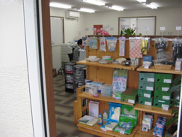 日用品棚と人物 エントランス 様々な日用品が揃うスペースでは、介護に必要なグッズを手軽に入手できます。奥にはスタッフが対応中です。