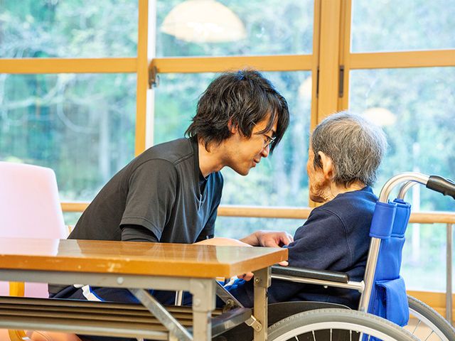 車椅子の高齢者と介護士の会話 ラウンジ 自然光が差し込む落ち着いた空間で、入居者とスタッフが対話を楽しんでいます。リラックスしながら働けます。