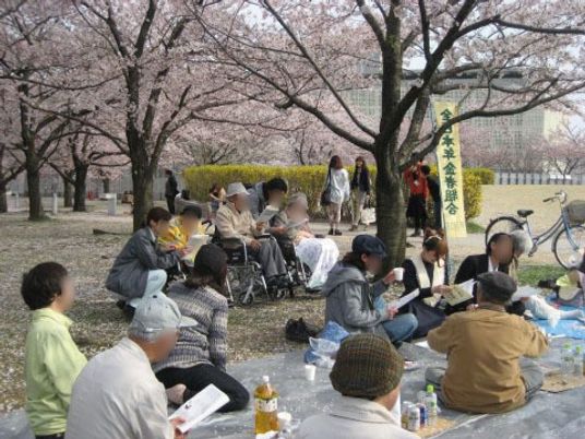 桜の咲く公園で、毎年恒例の施設の花見を行っている。入居者様と施設のスタッフが楽しい時間を過ごしている。