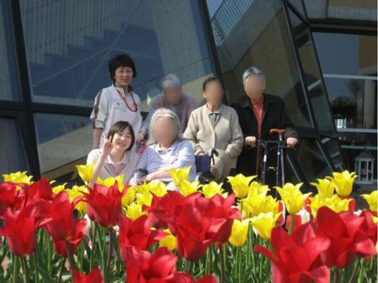 赤と黄色の美しいチューリップの咲く施設の中庭で、スタッフと入居者様が皆、笑顔で仲良く写真を撮っている。