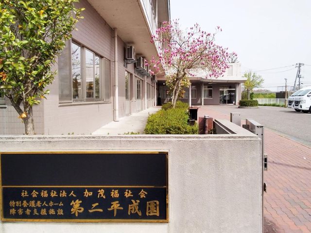 施設の看板と外観 看板 介護施設の名称が刻まれた看板と、その背後には緑が美しい施設の外観が広がっています。