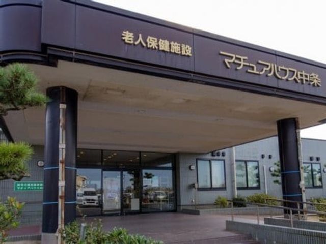 老人保健施設の入口 外観   落ち着いた色合いの外観に、ガラス張りの出入口が設けられています。手前にスロープも見え、出入りしやすそうです。