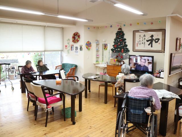 共有スペースでの日常風景 共有スペース ゆったりとした共有ラウンジで、利用者がテレビを観ながら過ごしています。椅子や車椅子でおくつろぎいただけるアットホームな空間が広がっています。