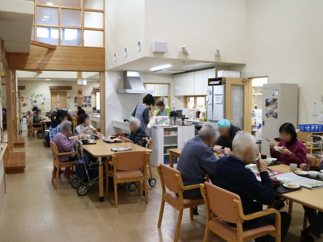 温かみのあるダイニング 共有スペース 明るく木造の食堂で、利用者が食事を取りながら談笑している光景です。スタッフも含めてコミュニケーションが活発に行われている様子が伺えます。