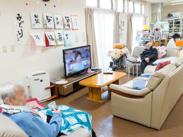 共有リビングの日常 共有スペース 柔らかい光が差し込むリビングで、利用者の方がくつろいでテレビを楽しんでいます。お気に入りの場所で日々のひとときをお過ごしいただけます。