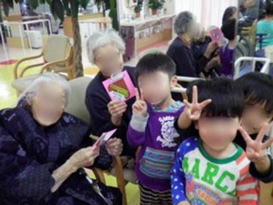 地域の子供たち３人と２人の入居者様が写っている。入居者様の手には子供たちからもらったカードが握られている。
