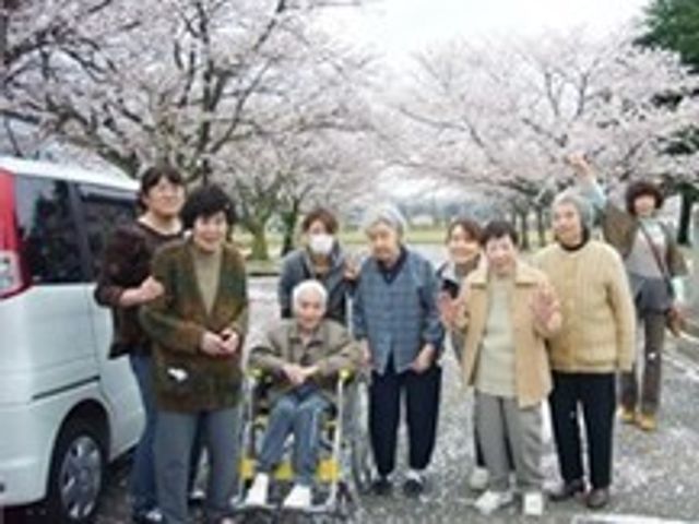 桜と笑顔の集合写真 外出レク 桜並木でのひと時を楽しむ利用者とスタッフ。和やかな雰囲気が伝わる場所で、共に過ごす時間があなたを待っています。