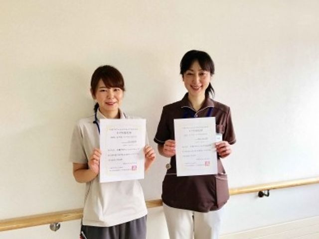 スタッフ2人の掲示前 掲示物   壁の前で2人のスタッフが書類を手に並んでいます。明るくすっきりした空間で、就業後の雰囲気を想像しやすい場面です。