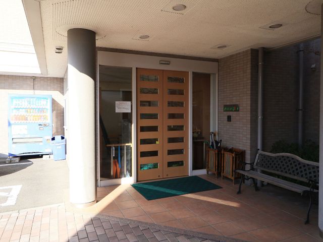 明るい施設のエントランス 玄関 落ち着いた色合いのエントランスには、待合のベンチがあり、訪れる人を温かく迎えます。周囲には植物が配され、心地よい雰囲気が漂っています。