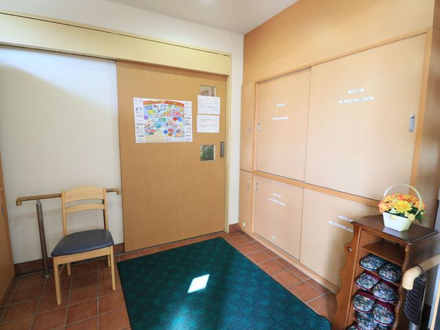 廊下の手すりと掲示物 玄関 椅子と鮮やかな花が置かれた廊下は、訪れる人の落ち着ける空間を提供しています。壁掛けの掲示物からは、日々の生活に役立つ情報を得られそうです。