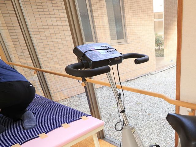 リハビリ機器と利用者 エアロバイク 明るい窓辺で、リハビリ器具を使用している方の姿が見られます。一人ひとりの体力に合わせた訓練ができる環境が整っています。