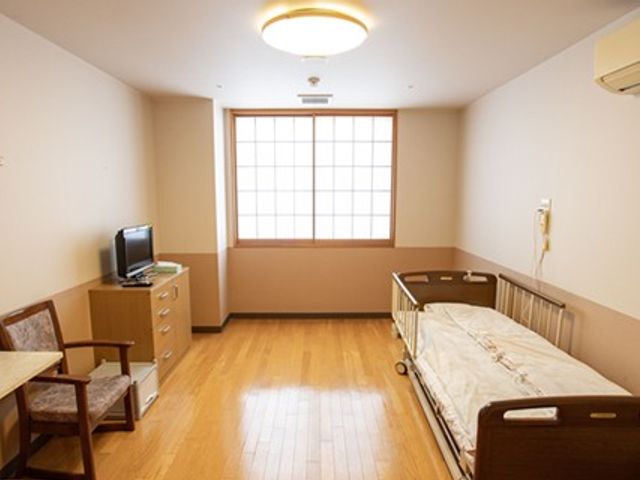 居室のベッドと窓 居室   木目調の床が広がる、明るく落ち着いた居室です。窓から自然光が入り、ベッドやテレビも見えます。