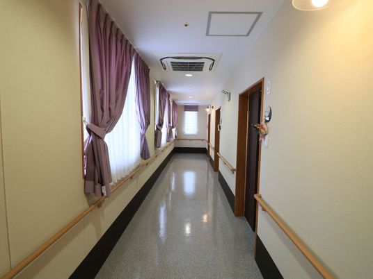 施設の写真 幅のある廊下には、障害物がなく、壁には手すりが設置されている。窓には、薄紫色のカーテンが掛かっている。