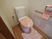 サムネイル 施設の写真 木目調の床に白い壁の清潔感のあるトイレである。便座には、ピンクの地に白い花柄が施されたカバーやマットが掛かっている。