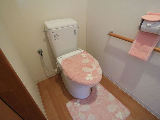 施設の写真 木目調の床に白い壁の清潔感のあるトイレである。便座には、ピンクの地に白い花柄が施されたカバーやマットが掛かっている。