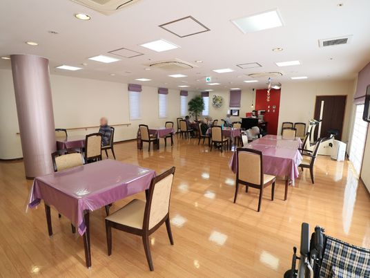 施設の写真 奥行きがあり、開放的な食堂である。薄紫のテーブルクロスにビニールカバーを掛けたテーブルと、椅子が配置されている。