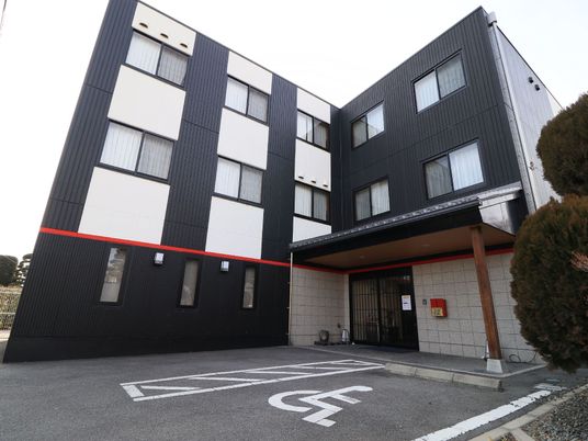 施設の写真 Ｌ字型をした３階建ての施設建物。コンクリート調の玄関周りと、黒ベースに白をはめた壁、赤いラインが特徴的である。