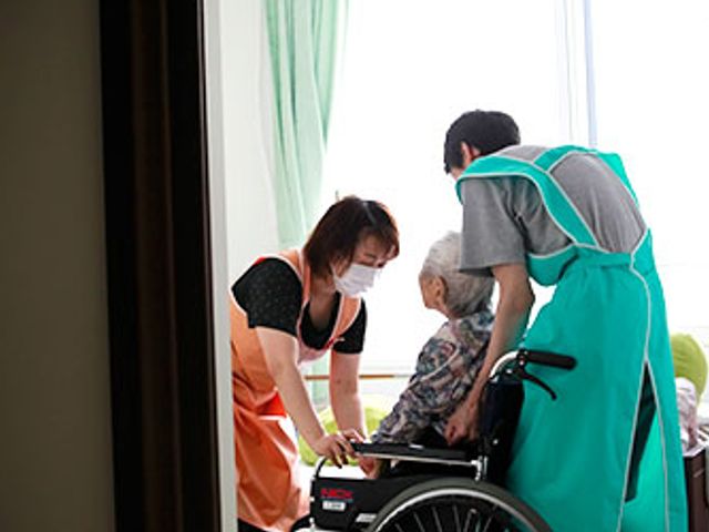スタッフと車椅子の様子 居室 スタッフが車椅子利用者をサポートしています。自然光が差し込む明るい空間です。