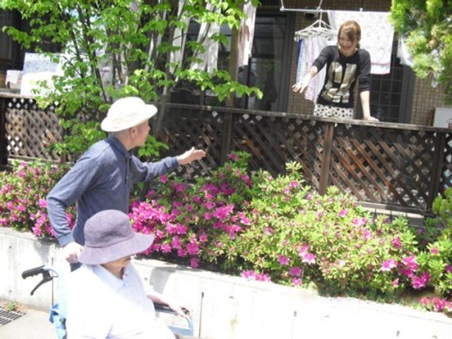 中庭の人々と植物 ケア風景 屋外で植物の手入れをするスタッフと、車椅子の方が交流している様子です。日差しを浴びながら、穏やかなひとときを過ごしておられます。