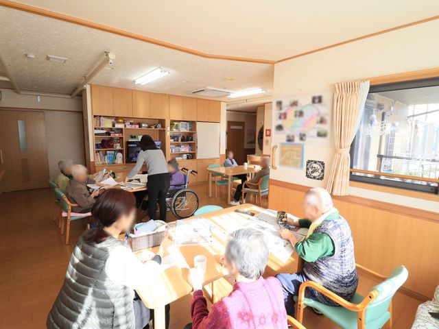 日光浴びる共有スペース 食堂 明るく穏やかな雰囲気のある空間で、利用者様がそれぞれの趣味に没頭されています。日差しの入る窓際では、手作業で楽しむ方々の姿が見受けられます。