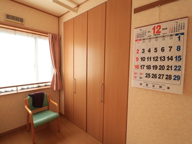 居室の椅子とカレンダー 収納 やわらかな光が差し込む居室で、壁に掲げられたカレンダーが日々の予定をお知らせします。窓から見える外の景色も気持ちを和ませてくれる空間です。