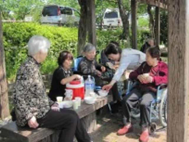 屋外でお茶する人々 スタッフ 木陰の涼しい中庭で、利用者の方々が憩いのひとときを過ごされています。明るい笑顔で心温まるケアを実感できる場所です。
