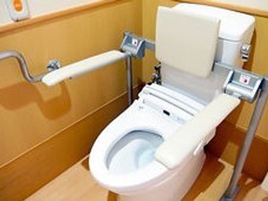 背もたれや手水が付いたトイレ。壁にはオレンジの呼び出しボタンも設置されている。