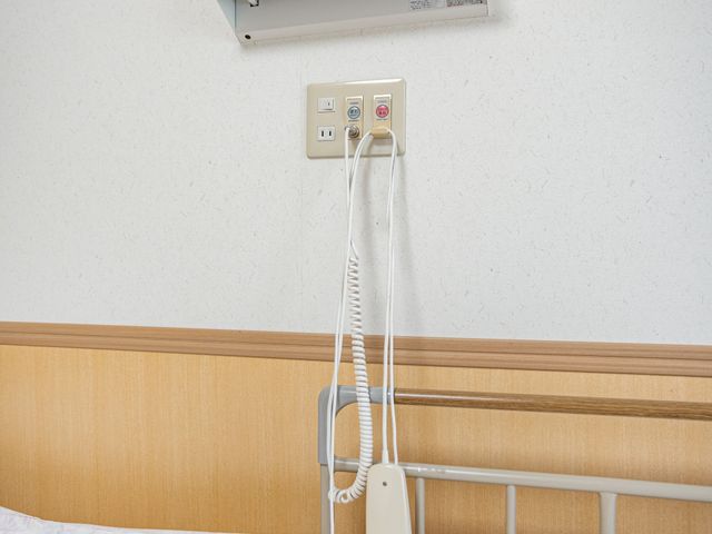 ベッド横の呼び出し装置居室 居室   ベッド横に呼び出し装置が備わっており、見守りや緊急時の対応がしやすい環境です。木目調の落ち着いた空間で、安心して過ごしやすそうです。