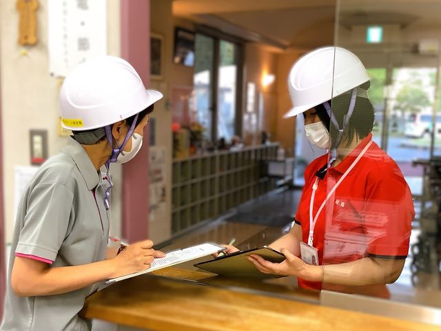 ヘルメットの職員が相談する受付 フロント   ヘルメットを着けた職員同士が、受付で書類を見ながら打ち合わせています。出入口近くで来訪者対応の流れを確認する様子がうかがえます。