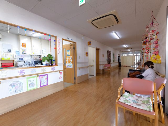 暖かみのある廊下の風景 廊下 座ってくつろぐ人がいる廊下は心地よい雰囲気で、季節の飾りつけも楽しめるスペースです。利用者の笑顔が印象的な、あたたかな通り抜けスペースとなっています。