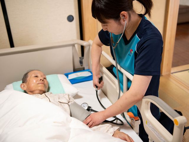 健康管理のケアシーン 優しい手つきで血圧を測るスタッフと、安心して委ねる利用者の様子です。ケアの質の高さを感じられます。