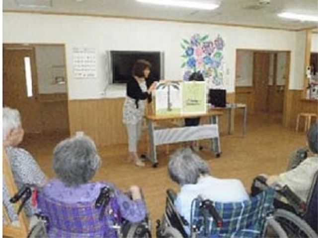 多目的スペースの一コマ 紙芝居 ゆったりとした多目的室で、利用者が集う暖かい雰囲気です。スタッフが心地よい対応をしている様子が伝わります。