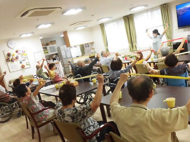 運動を楽しむ人々の居室 居室 自然光が差し込む広々とした居室で、参加者たちはストレッチの運動を楽しんでいます。車椅子の方も一緒に参加できる環境です。