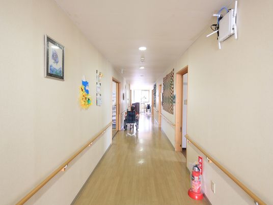 施設の写真 白い壁と明るいベージュの床の廊下がある。壁にはカラフルな布や絵が飾られ、手すりがある。床には消火器が置かれている。