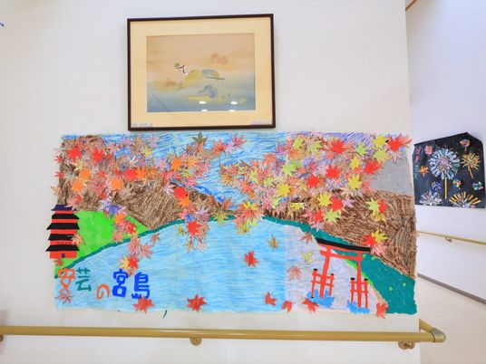 廊下の壁に、貼り絵などで作られた絵画作品が飾られている。手前は宮島の風景で、奥には花火の柄のものが見える。