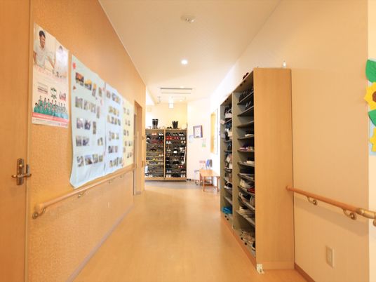 施設の写真 通路右側と正面に背の高い靴箱が各２つ置かれている。壁には手すりが取り付けられており、その上にポスターや入居者様の写真が貼られている。