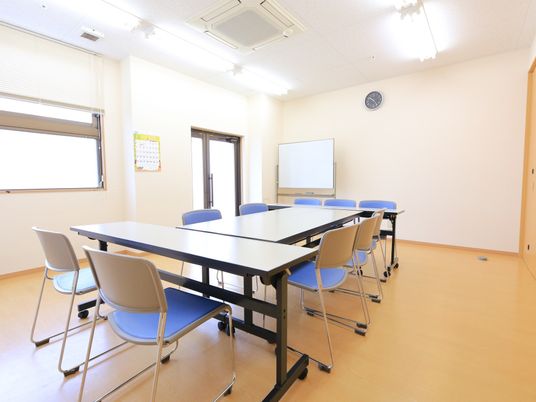 白い壁とライトブラウンの床の共有スペースがある。大きな机と椅子がある。ホワイトボードがあり、壁には時計が掛かっている。