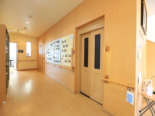 施設の写真 フローリングでベージュ色の壁の通路がある。壁には手すりが備え付けられており、模造紙に貼られた写真が掲示されている。